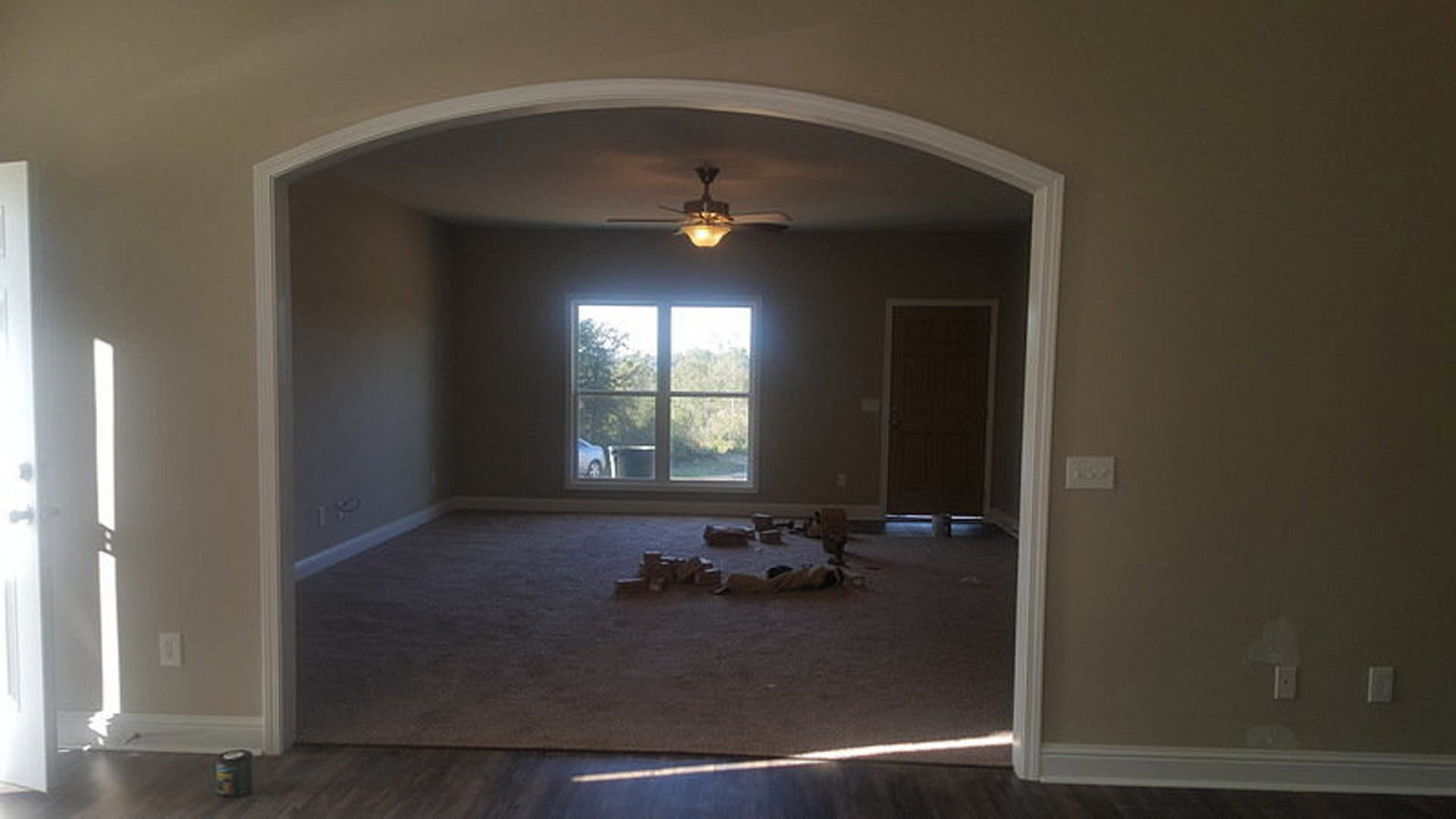 Living room with laminate flooring, ceiling fan, dark door with white frame, large window showing trees outside, plaster walls