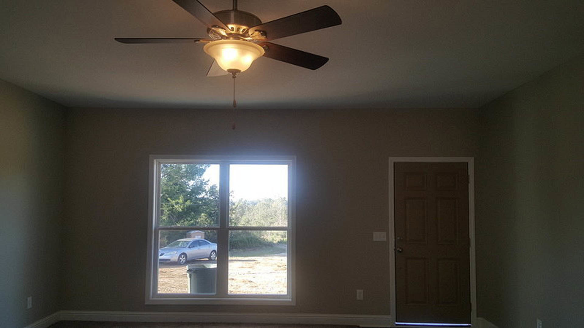 Ceiling fan with dark blades mounted on white living room ceiling, large windows, neutral walls, and recessed lighting visible.