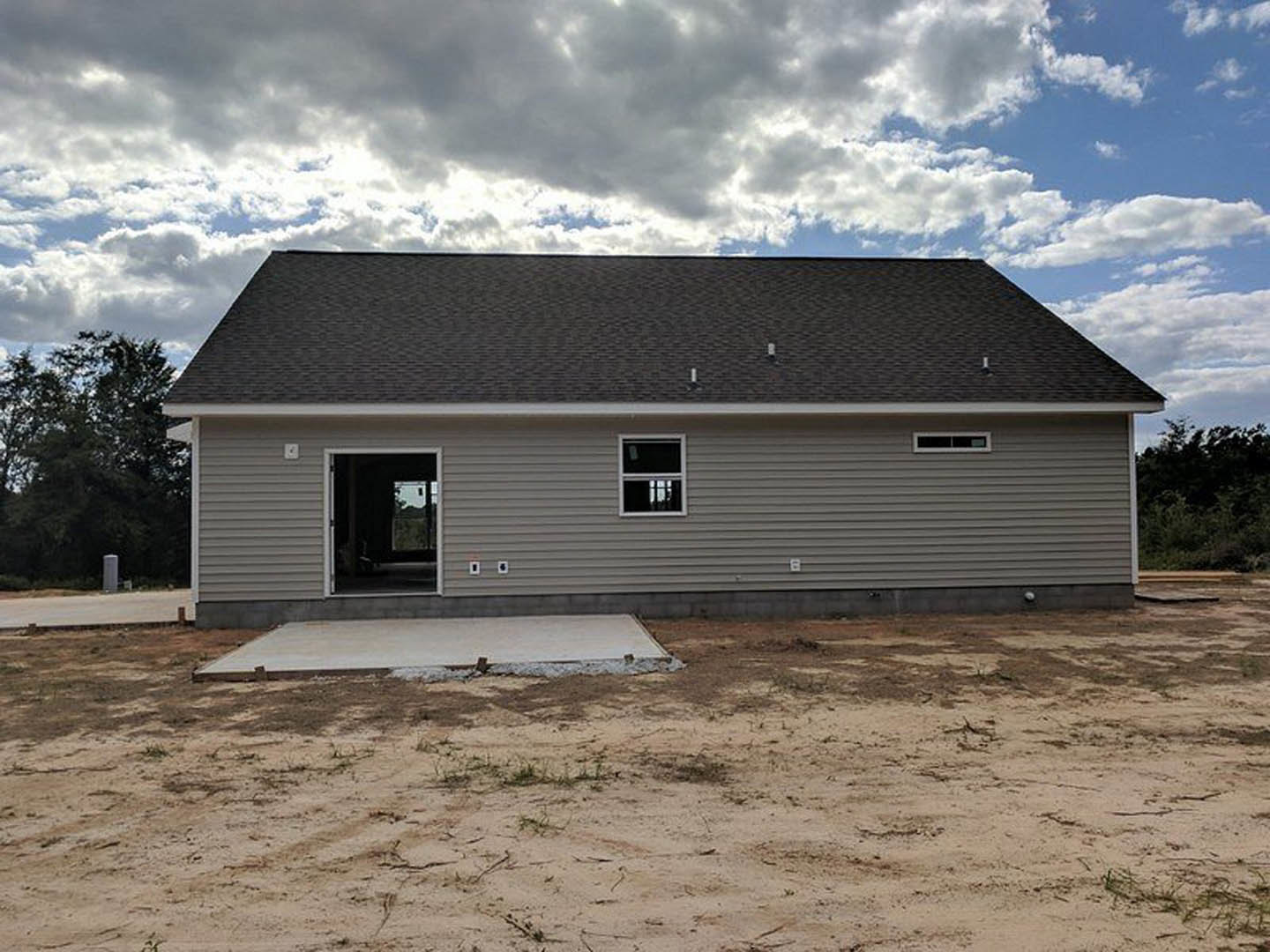 Partially built house with exposed concrete slab foundation, framed window opening, and unfinished siding under overcast sky, surrounded by dirt and sparse trees.