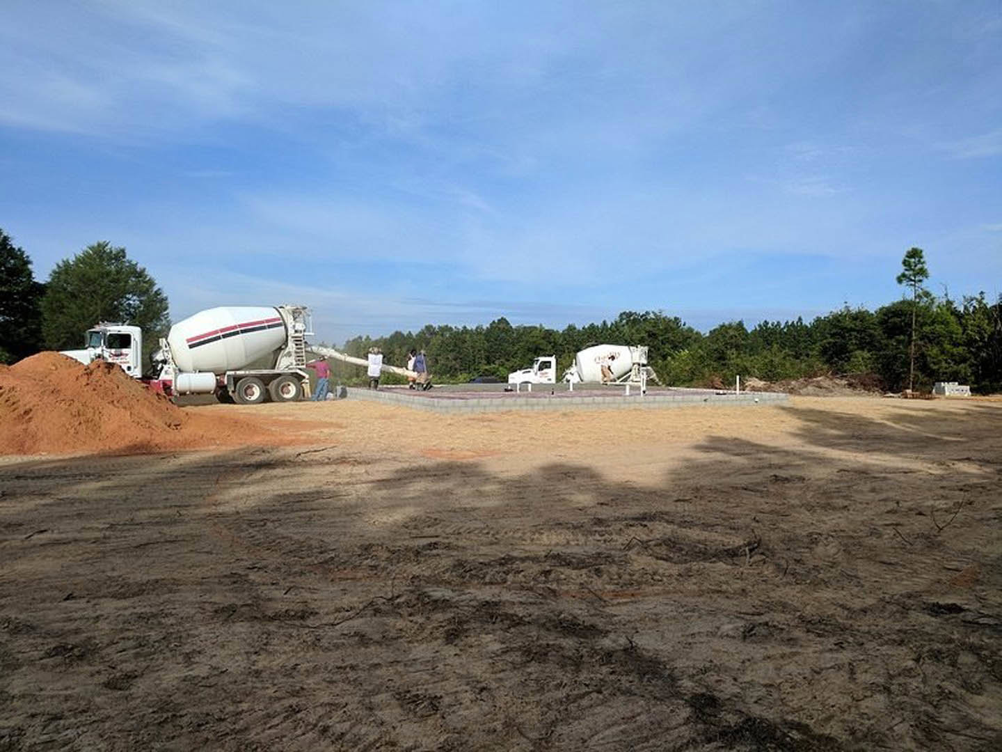 Cluster of white cement mixer trucks parked on dirt construction site under blue sky, with piles of soil and tree shadows visible.