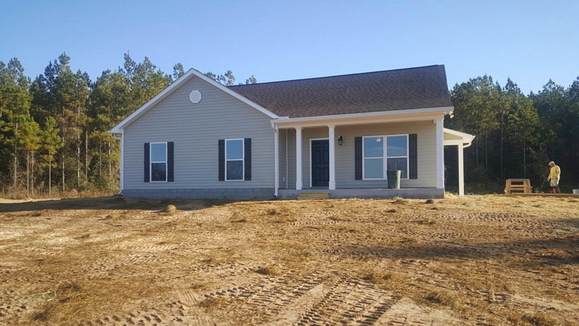Modern house with black front door, white-framed window, gray exterior wall, dirt field with tire tracks, white circular marking, and a tree in the background; blurry man in yellow