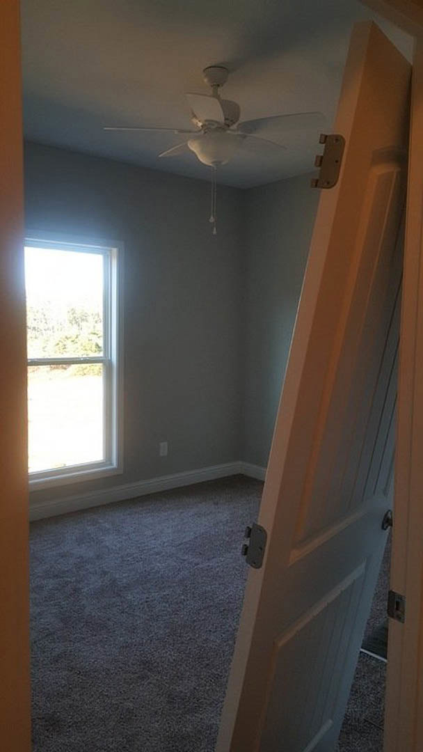 Open door leading to carpeted room with white walls, ceiling fan, wood post detail, and window overlooking trees