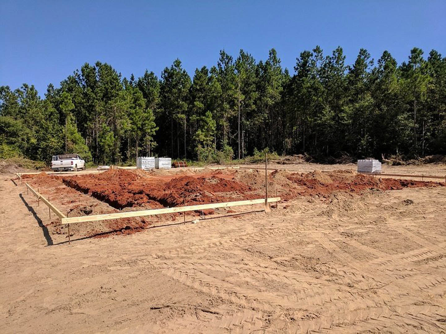 Dirt clearing with tire tracks, wooden fence, white truck parked, dense forest of trees in background under blue sky