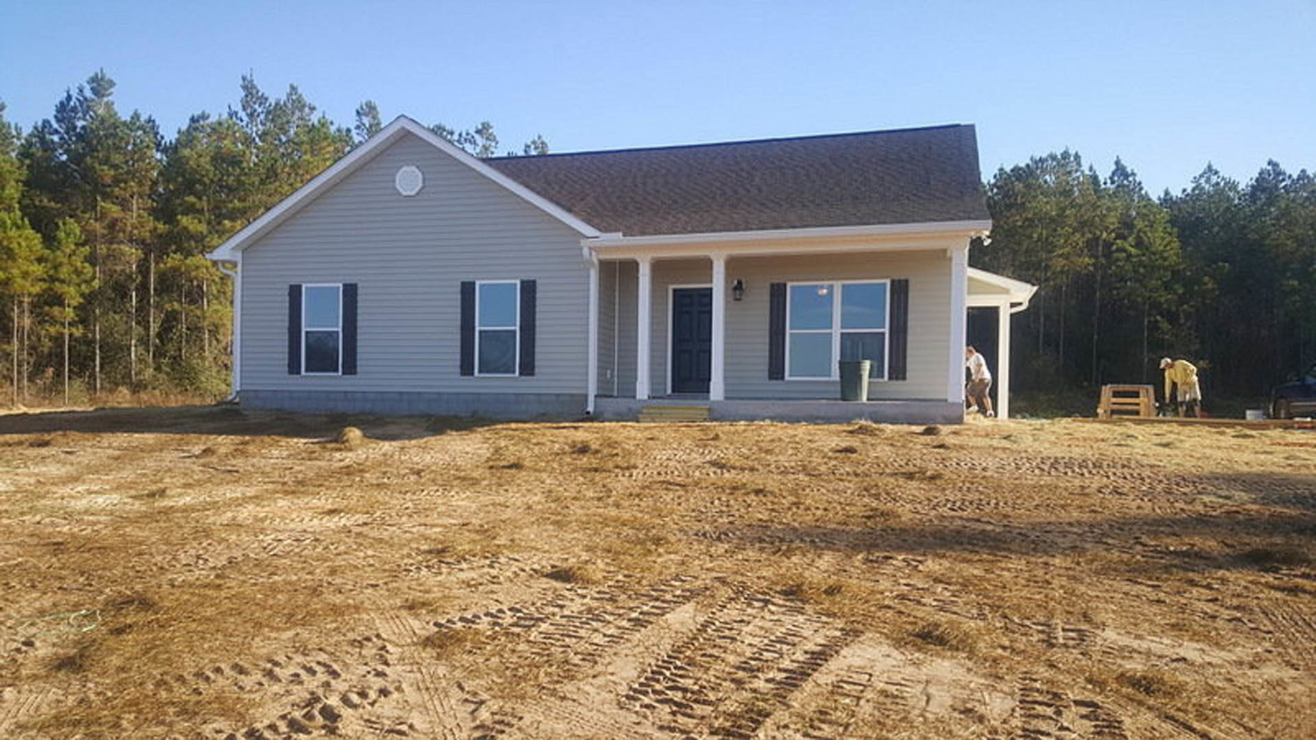Modern home exterior with black door, white-framed windows featuring square patterns, tire tracks on dirt lot, surrounded by trees