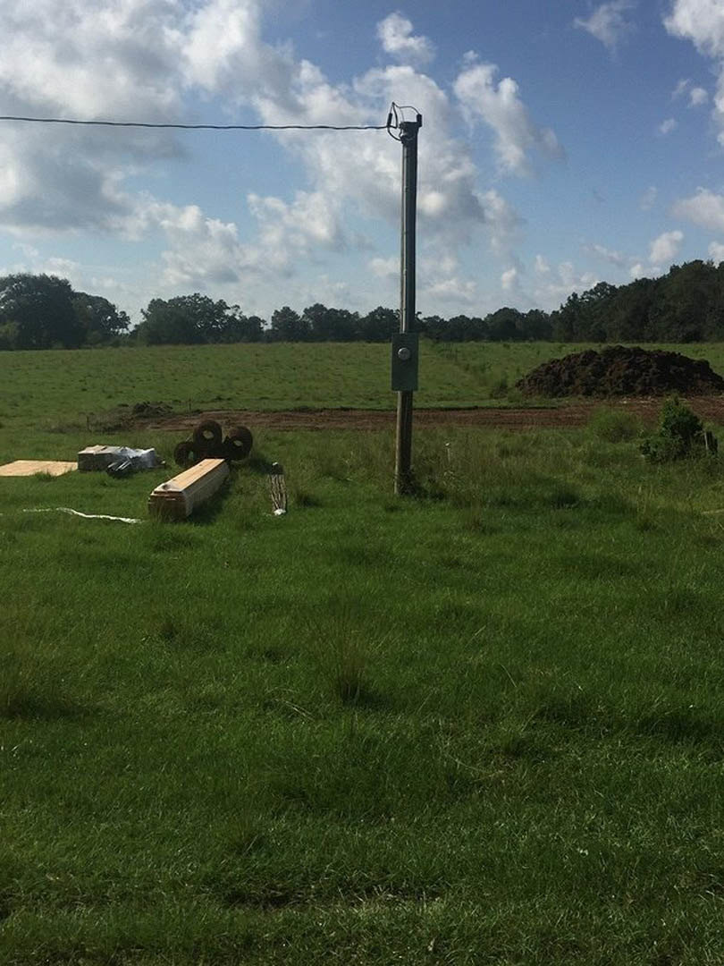 Power line pole with wood beam and coiled wire beside grassy field, wooden box and dirt pile in foreground, metal pole with light, fence, log, and scattered trees under cloudy sky.