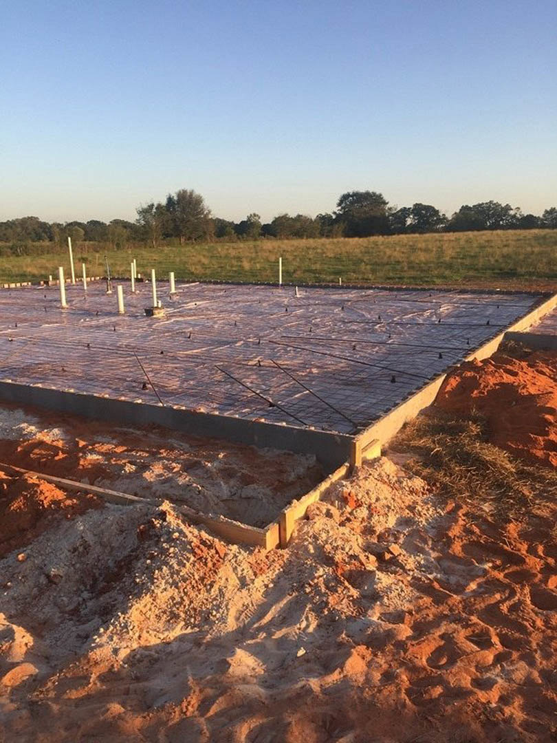 Concrete house foundation with plastic tarp covering, surrounded by grassy field and trees under partly cloudy blue sky