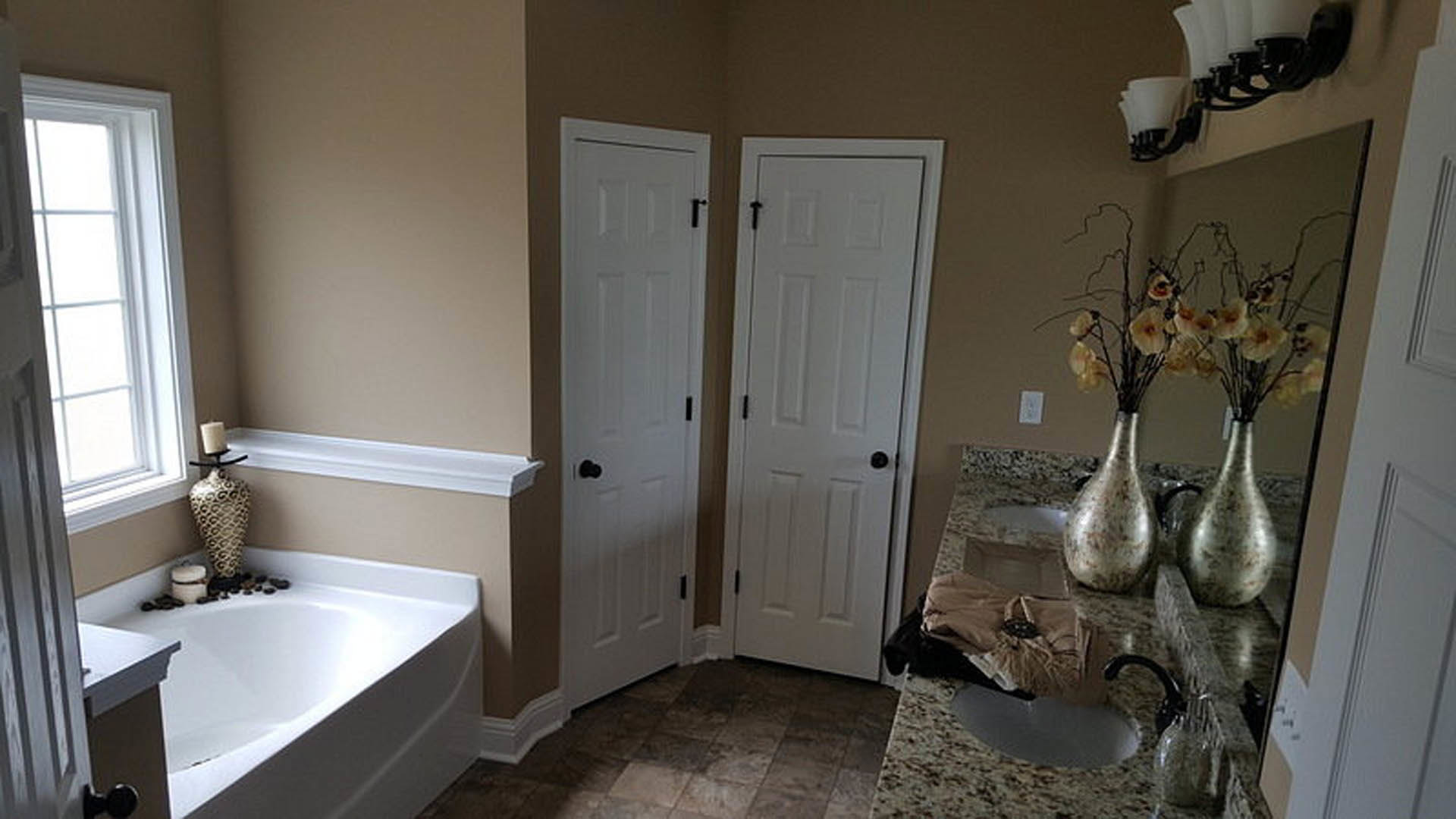 Bathroom with two white doors, freestanding bathtub beside decorative rocks, wall-mounted mirror above sink, neutral tile flooring, chrome faucet, and blurred vase in foreground