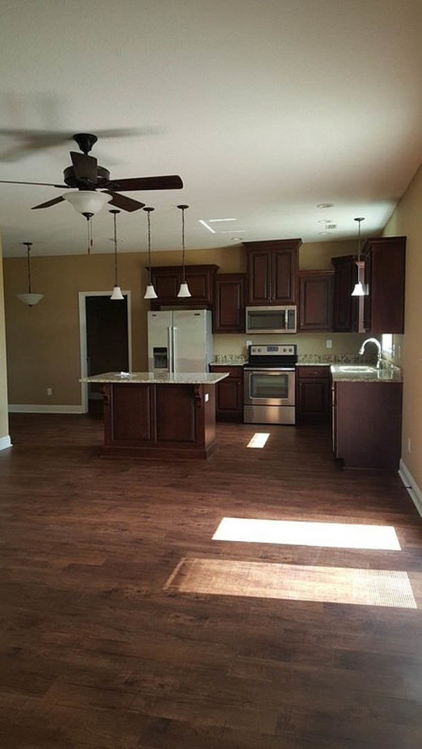 Kitchen with marble countertop, white refrigerator, built-in microwave, wooden flooring illuminated by sunlight, ceiling fan, and white rectangular object on counter.