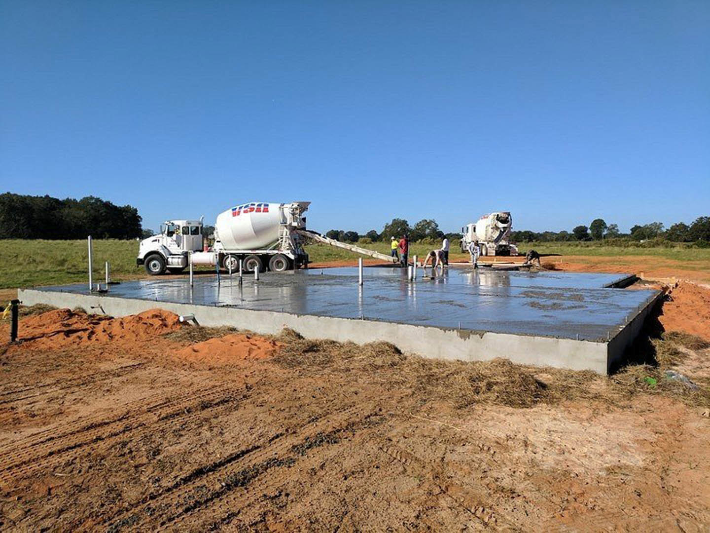 Concrete mixer truck parked beside freshly poured foundation, dirt field, hay pile, and partially built concrete wall; trees and blue sky in background.