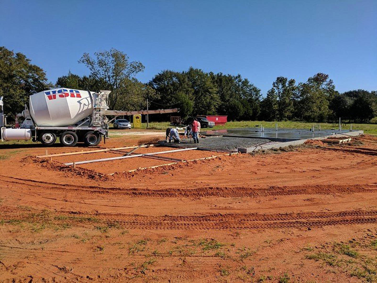 Cement mixer on trailer and construction truck parked on dirt lot, workers standing near unfinished foundation, surrounded by soil, grass, and trees under open sky