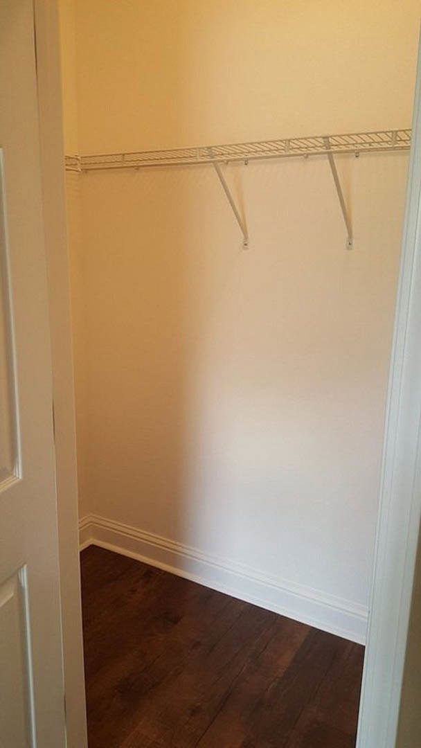 Closet with white plaster wall, wood floor, white shelf above metal rods for hanging clothes