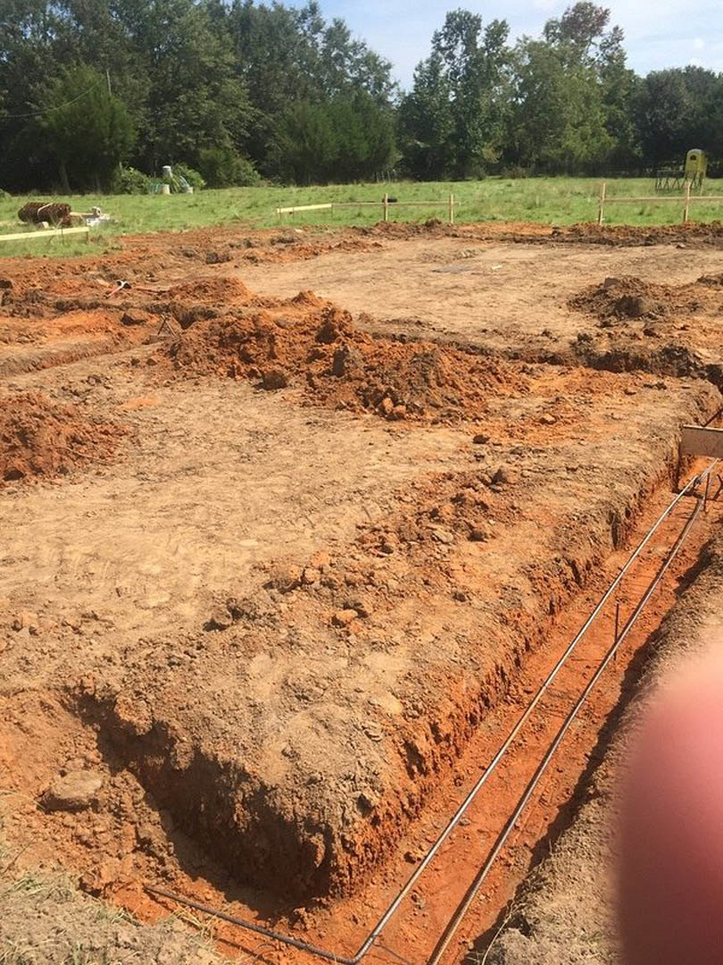 Dirt field with scattered metal pipes, patches of grass, and trees in the background under a cloudy sky
