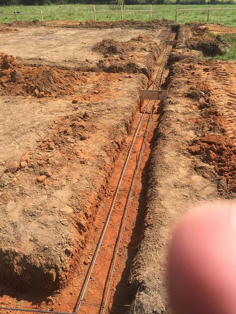 Metal pipes laid in a dirt trench surrounded by soil and grass, with a fence visible in the background.