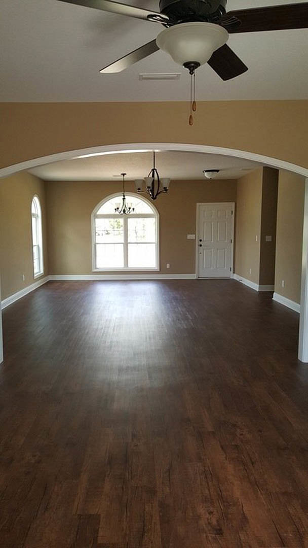 Wood flooring in a spacious room with a modern chandelier, white walls, white door with black hardware, and ceiling fan with light fixture.