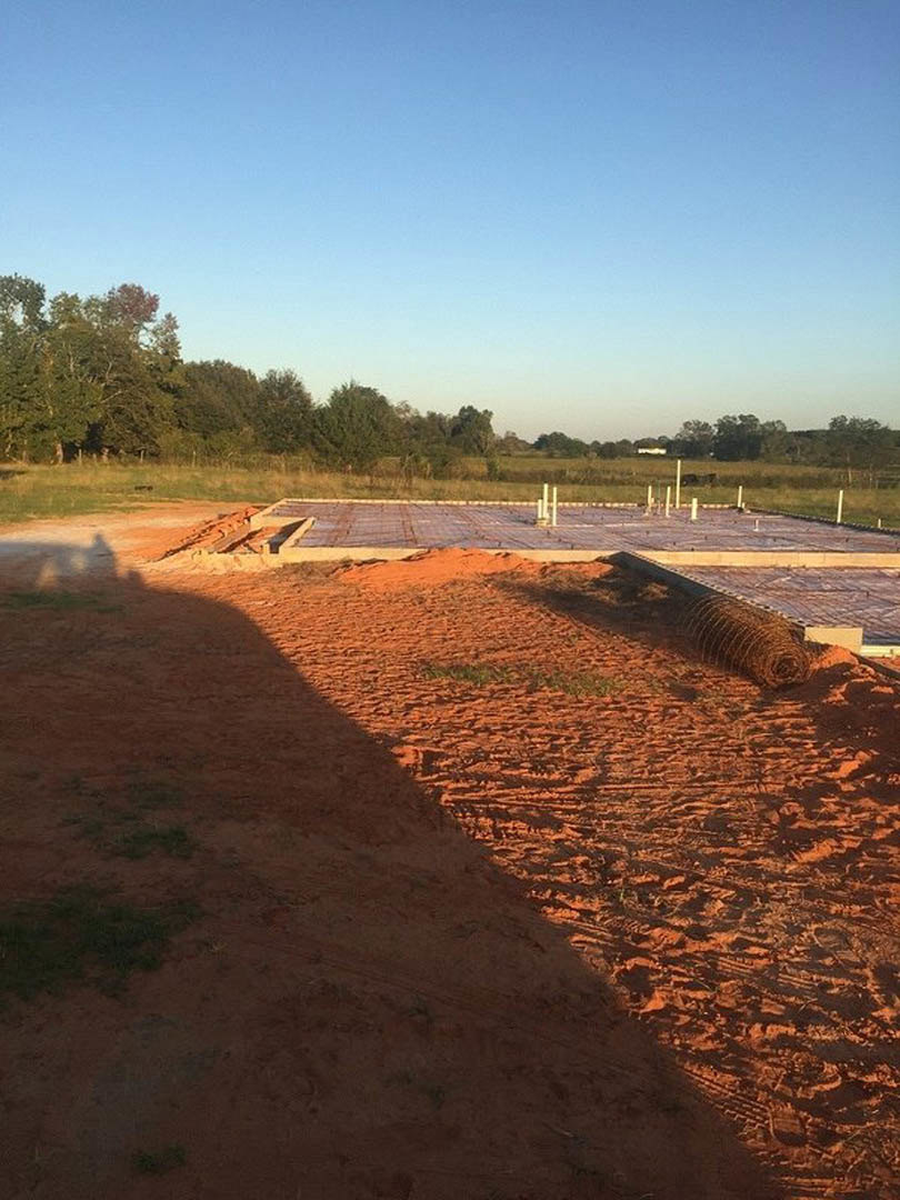 Metal roll resting on dirt ground at residential construction site, temporary fencing nearby, grassy field and trees under partly cloudy blue sky