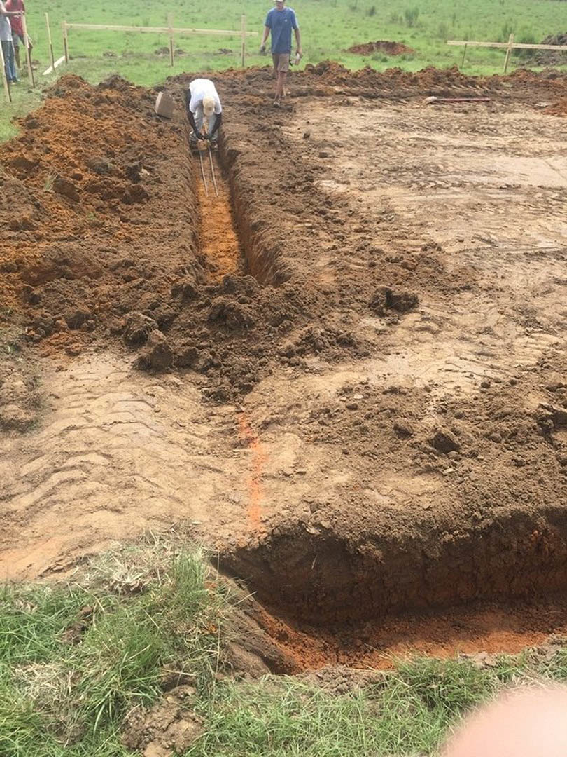 Man in blue shirt digging trench in muddy soil beside grassy field, surrounded by plants and dirt.