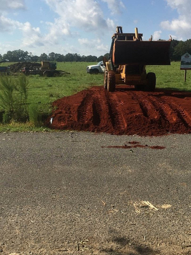 Tractor parked on a dirt road beside grassy land lot, tire tracks visible in soil, white truck and construction sign in background, trees and cloudy sky overhead