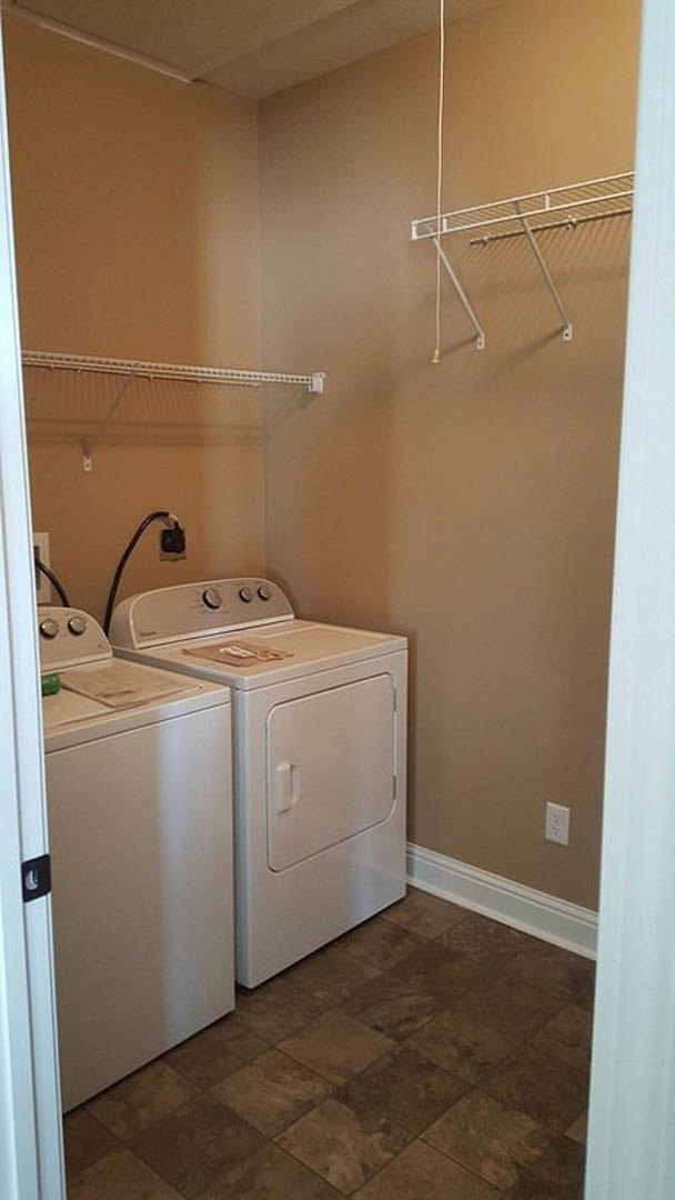 Front-loading washer and dryer set against a white wall in a laundry room with brown tile flooring, white cabinetry, and a metal hanging rod.