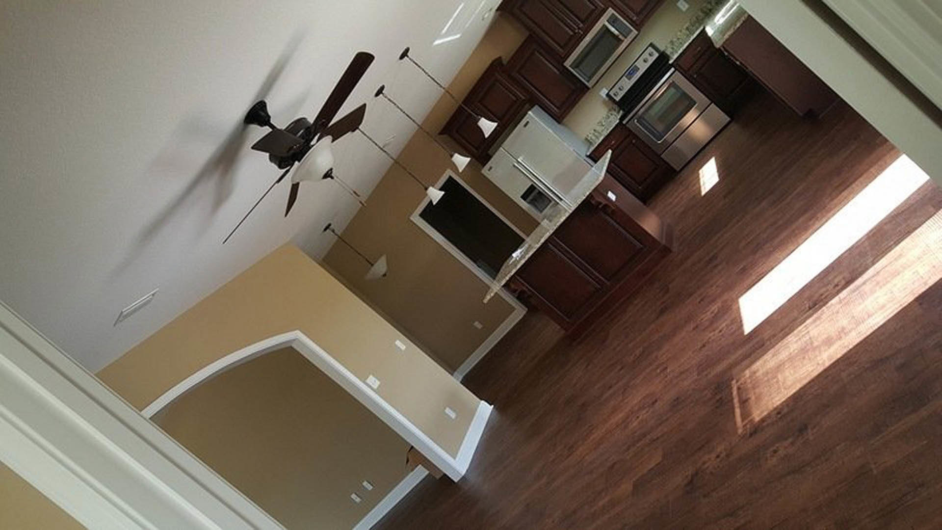 Kitchen with wood flooring, ceiling fan with light, black-framed window, white walls, hallway visible, built-in microwave, natural light streaming through window