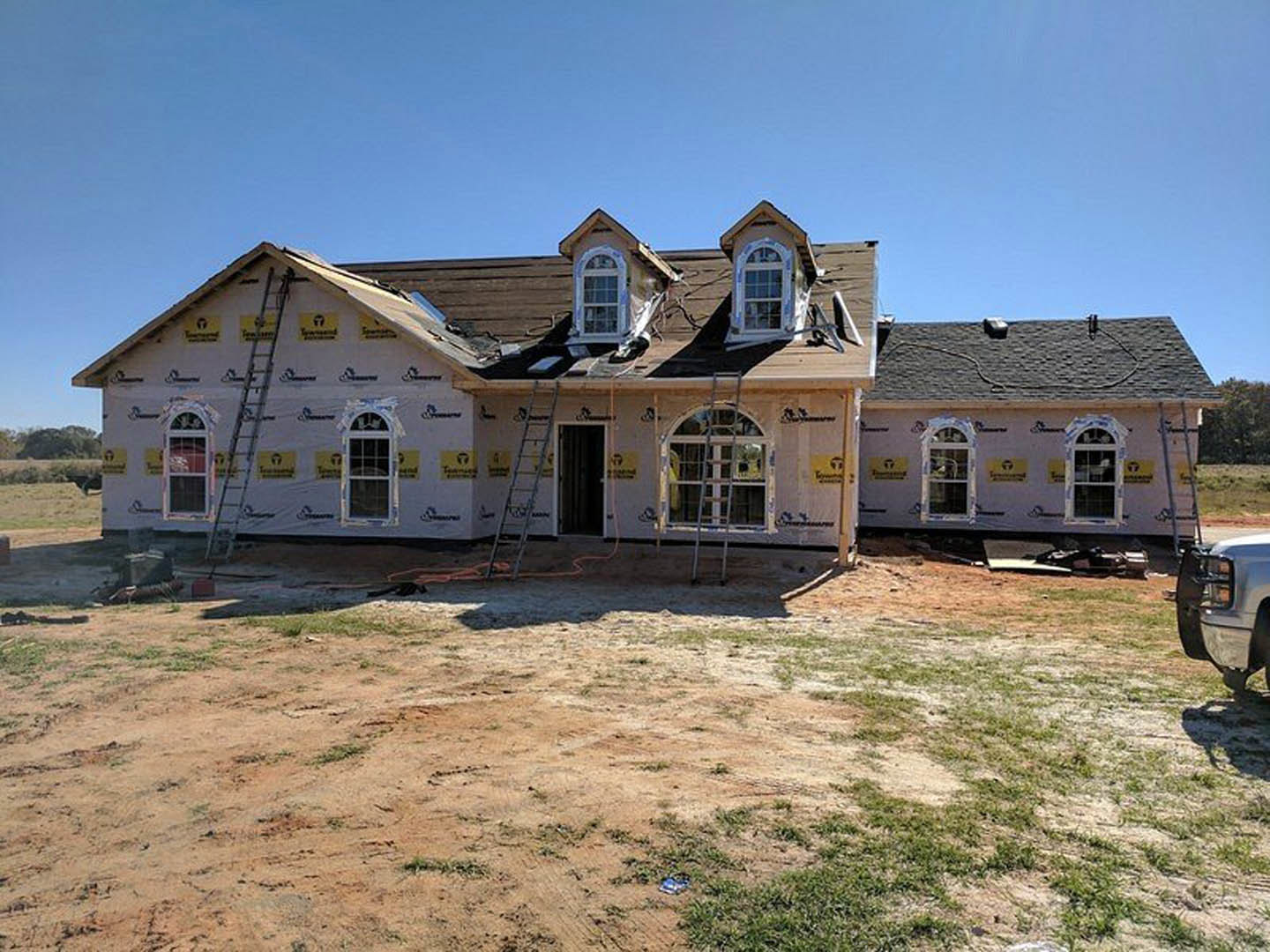 Framed house under construction with exposed wood, ladders leaning against exterior walls, dirt yard, unfinished windows, and construction materials scattered around