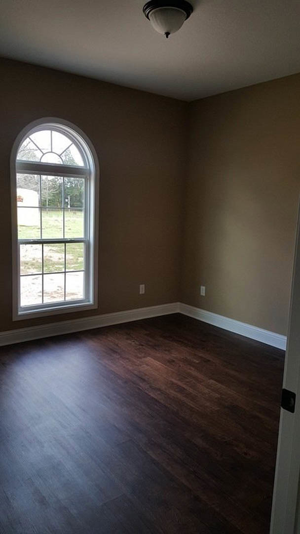 Corner of a room featuring a large window overlooking a grassy field, dark wood laminate flooring, white walls, and a ceiling light fixture.