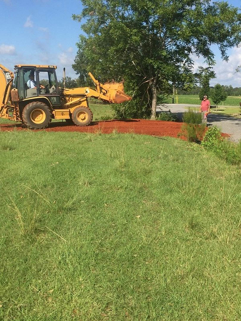 Green tractor with large black tires parked on a dirt driveway beside a grassy lawn, mature trees in background, blue sky overhead