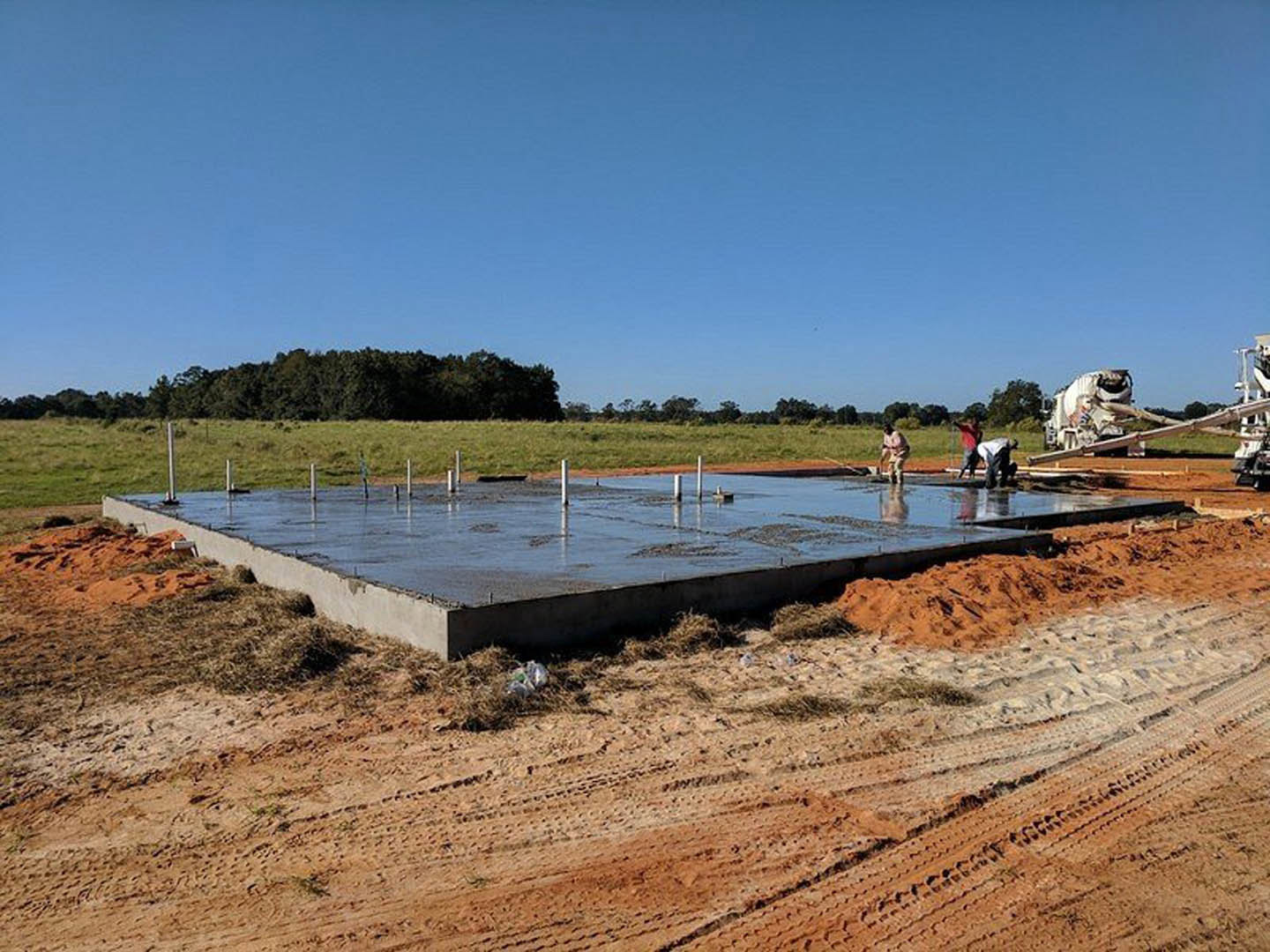 Concrete foundation slab with vertical poles at a residential construction site, grassy field and trees in background, blue sky with scattered clouds overhead, blurry figure of a