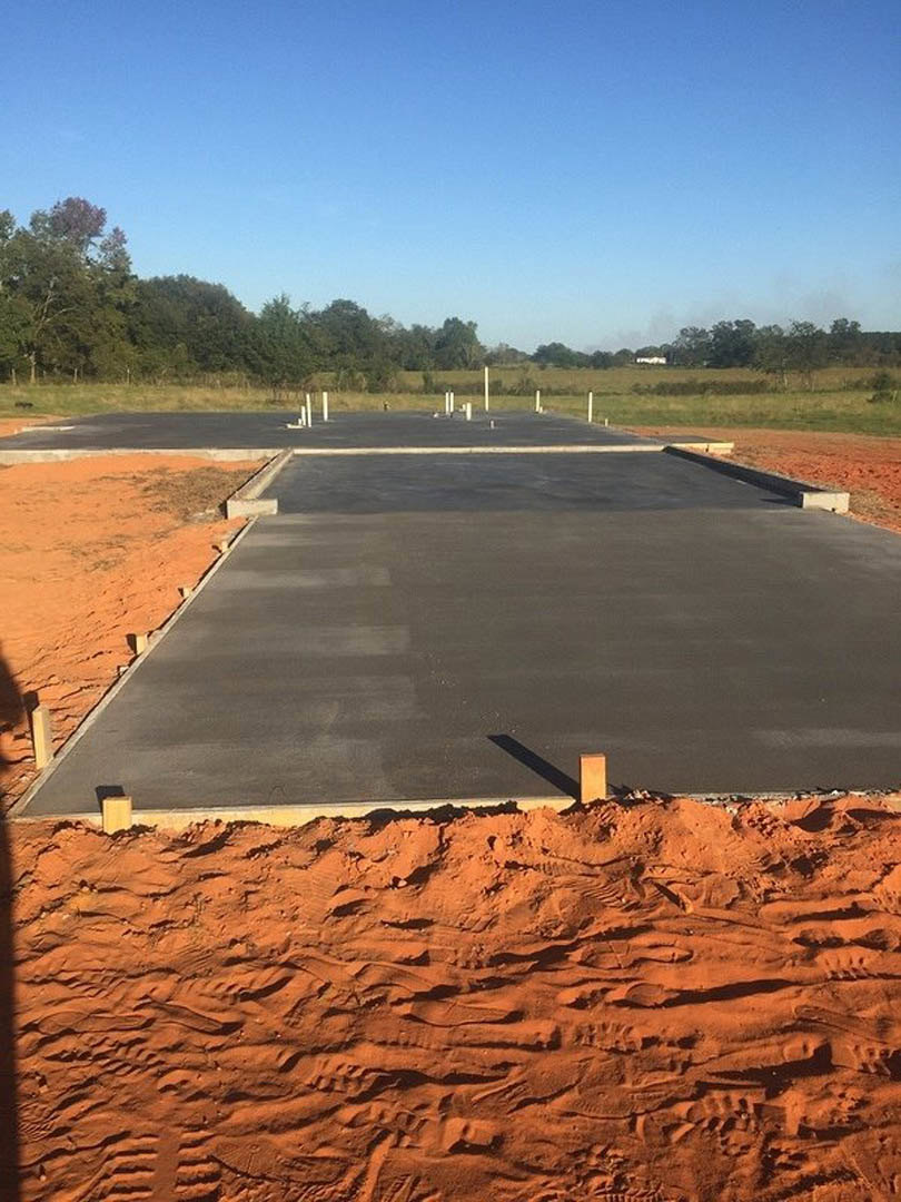 Concrete slab foundation under construction with metal poles, sandy ground, grassy field, and trees beneath a clear blue sky