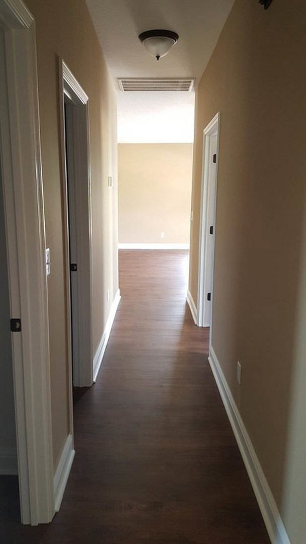 Hallway with smooth white walls, dark wood flooring, white door featuring a black handle, ceiling vent, and black trim along the wall