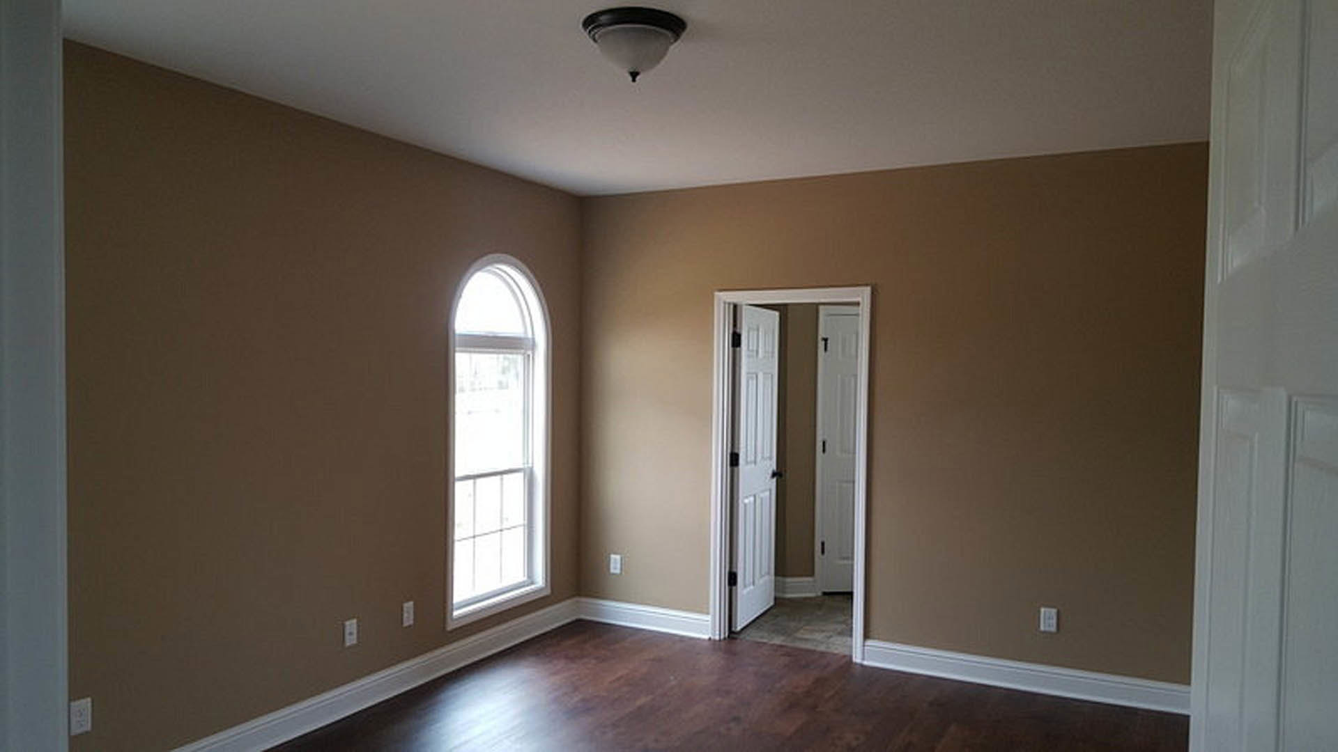 White paneled door open to a room with brown hardwood floors, white baseboard trim, and a window letting in natural light; modern light fixture visible on ceiling.