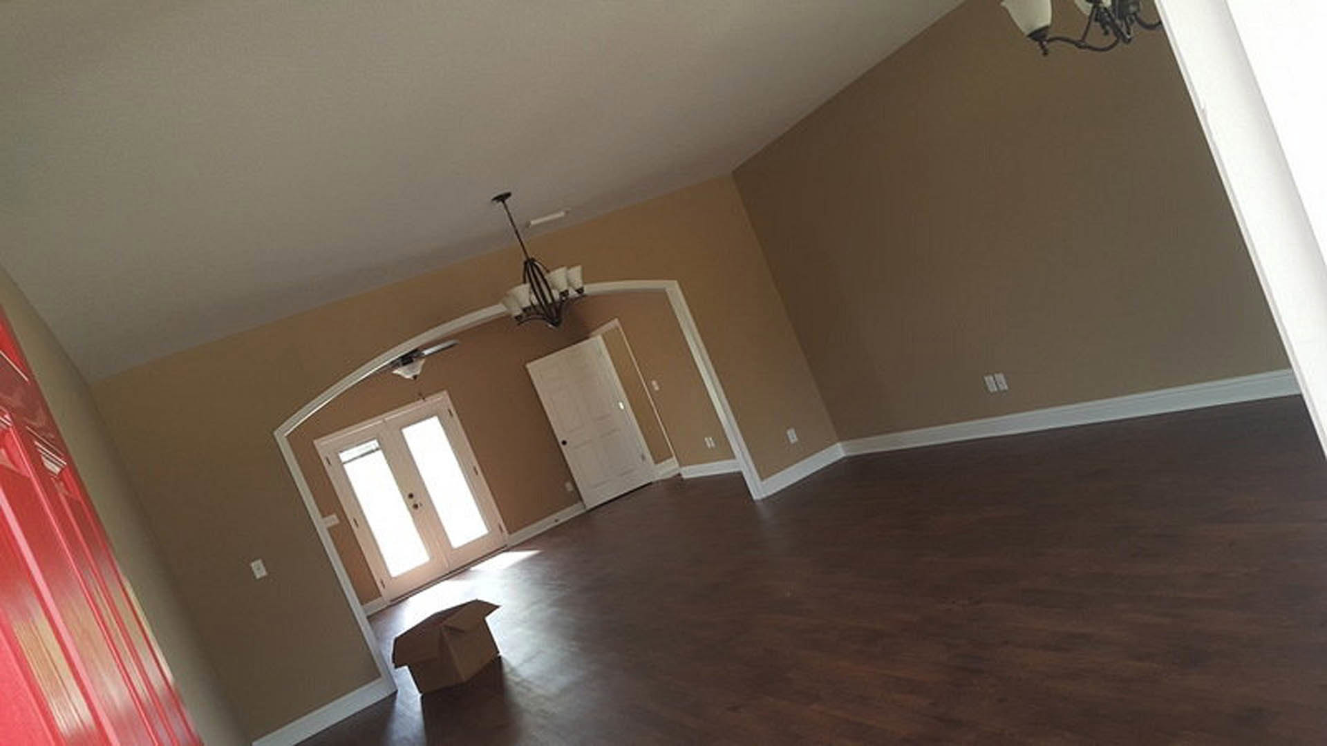 Open room with light wood flooring, white double doors with black knobs, white walls with crown molding, and a cardboard box placed near the center; chandelier partially visible