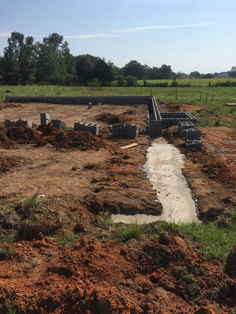 Concrete foundation set in a grassy field, surrounded by dirt piles, fencing, and open sky with scattered clouds