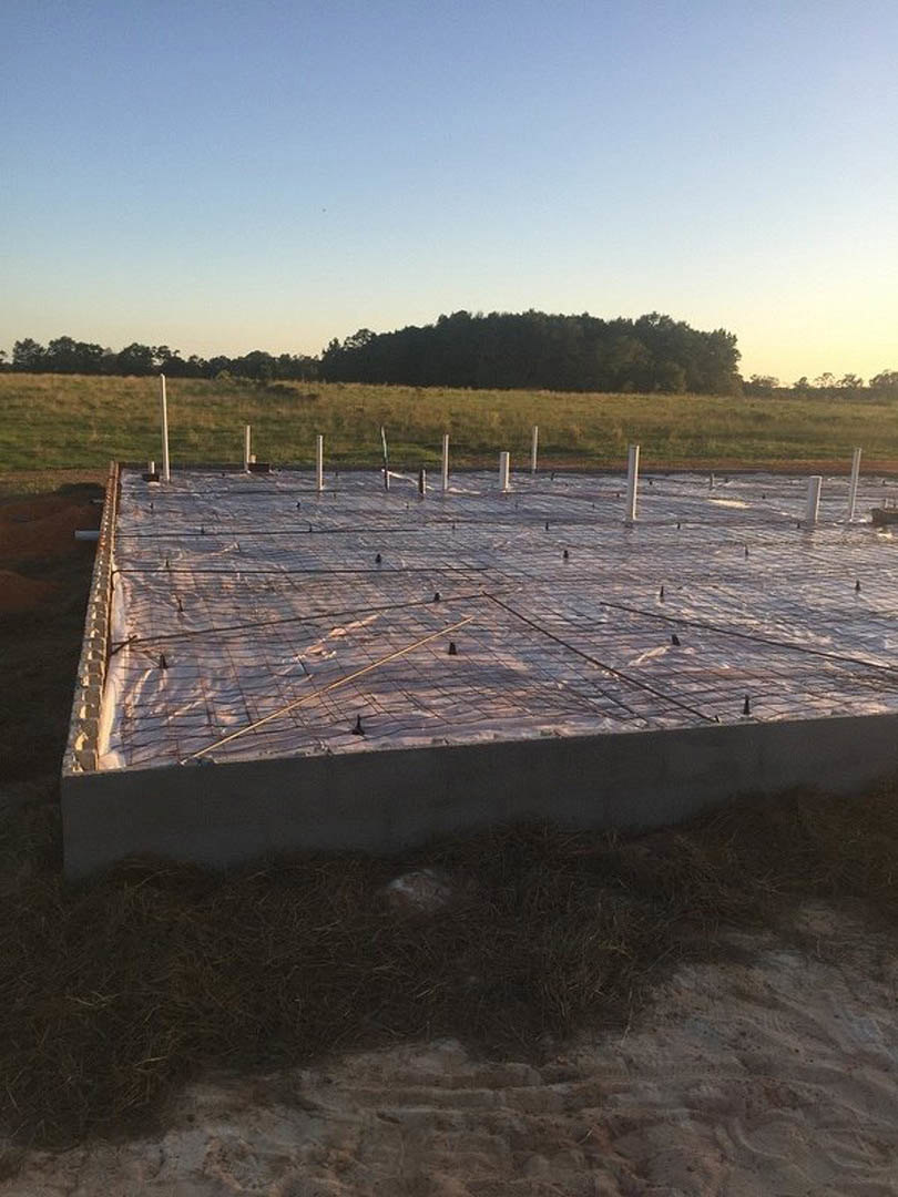 Concrete foundation with exposed metal rods, hay pile nearby, grassy field bordered by a fence, distant trees, and partly cloudy blue sky
