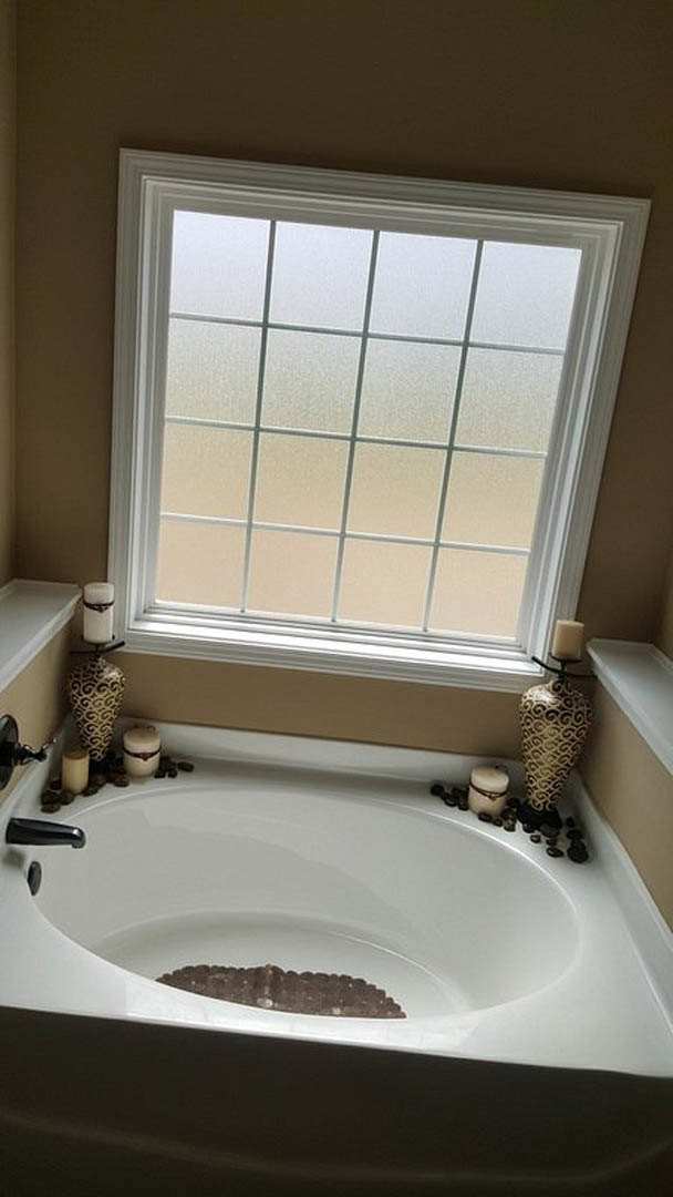 Freestanding white bathtub with lit candles on the rim, large window with natural light, neutral bathroom walls, chrome faucet, and decorative vase nearby