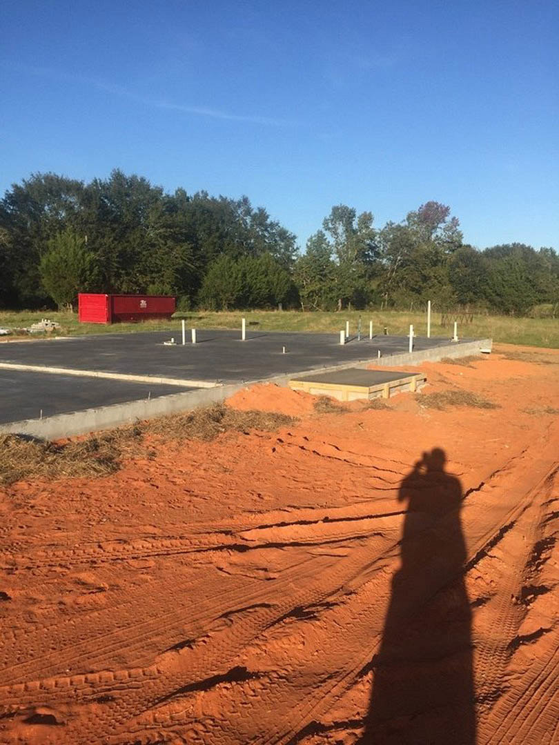 Shadow of a person cast on a dirt road bordered by trees under a blue sky with scattered clouds; red metal container with white writing visible in the background.
