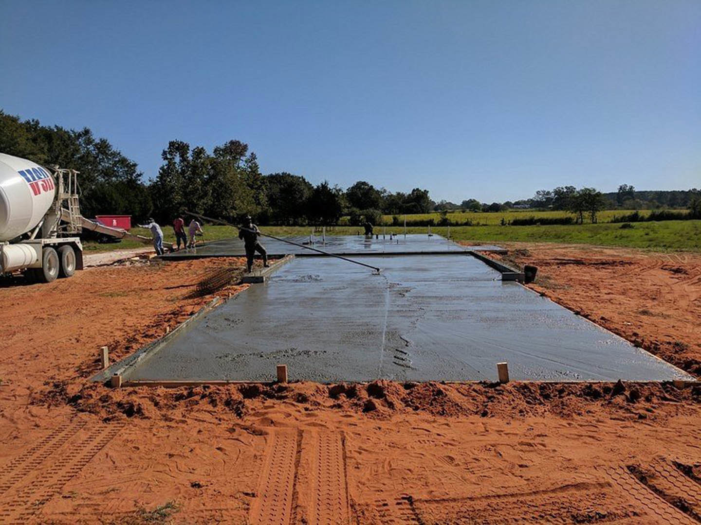 Concrete foundation slab surrounded by construction workers, cement mixer, and truck, with blue sky and trees in background