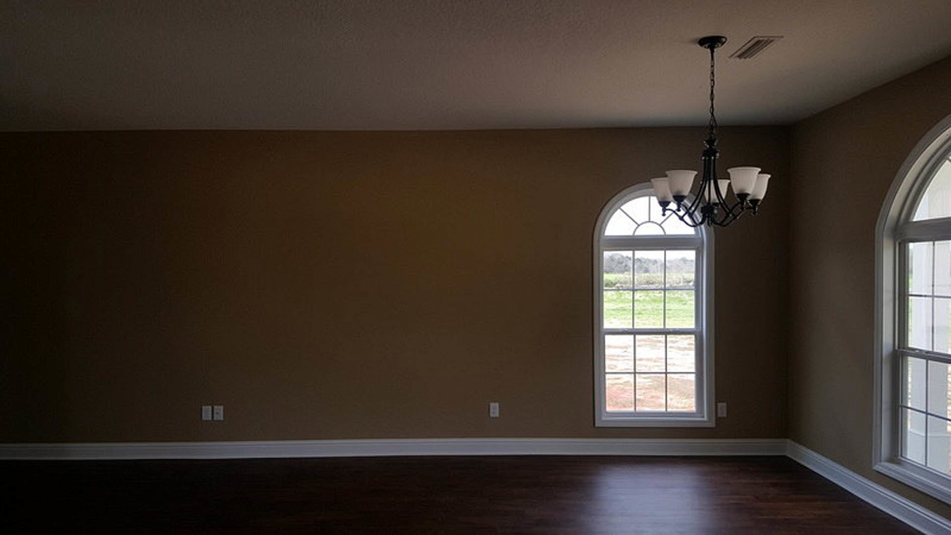 Crystal chandelier hanging from white plaster ceiling with crown molding, large window with grass field view, vent and window blinds visible