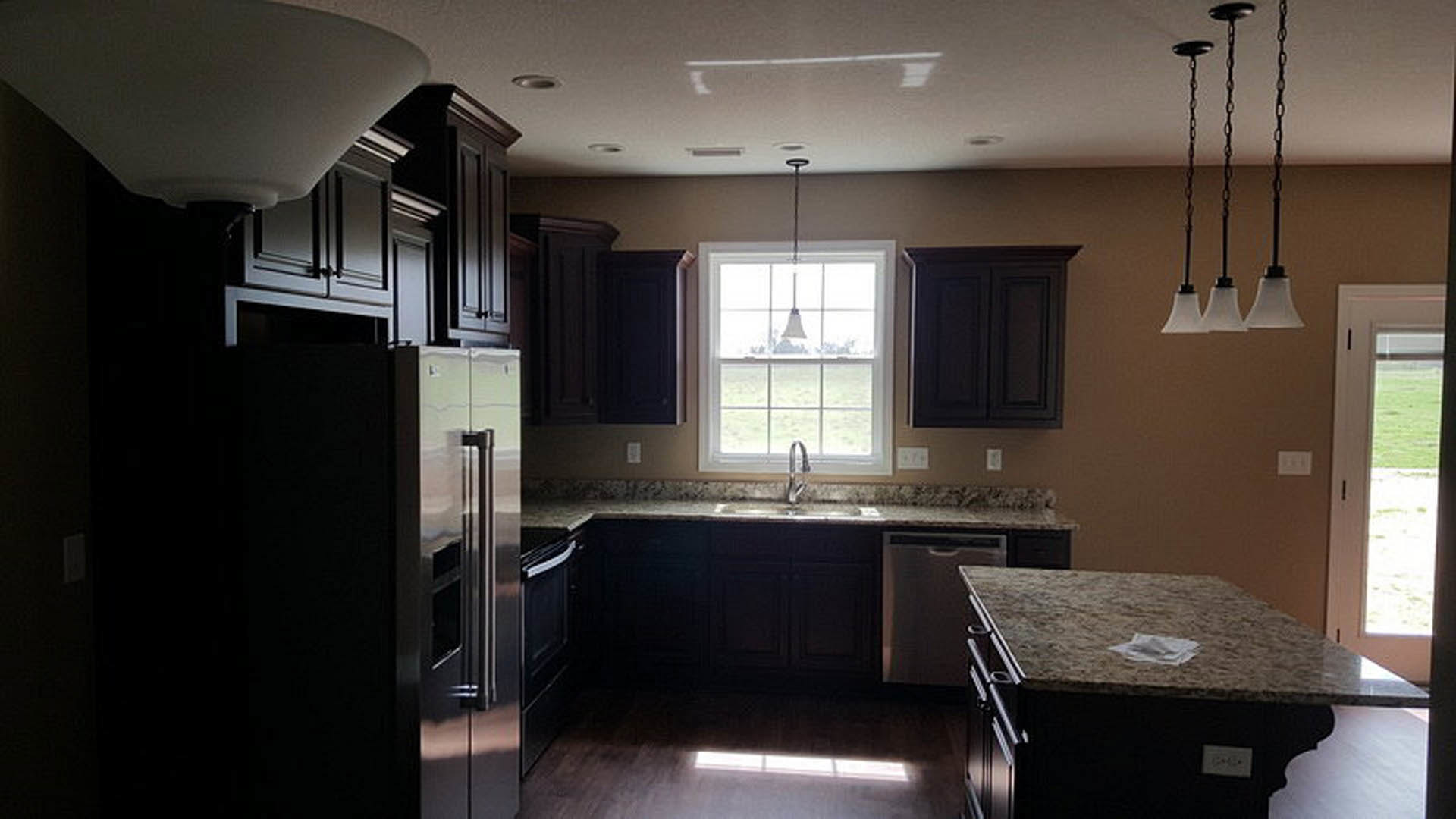Dark wood kitchen cabinets, stainless steel refrigerator, light stone countertop with a folded napkin, black rectangular appliance, undermount sink, and neutral walls.