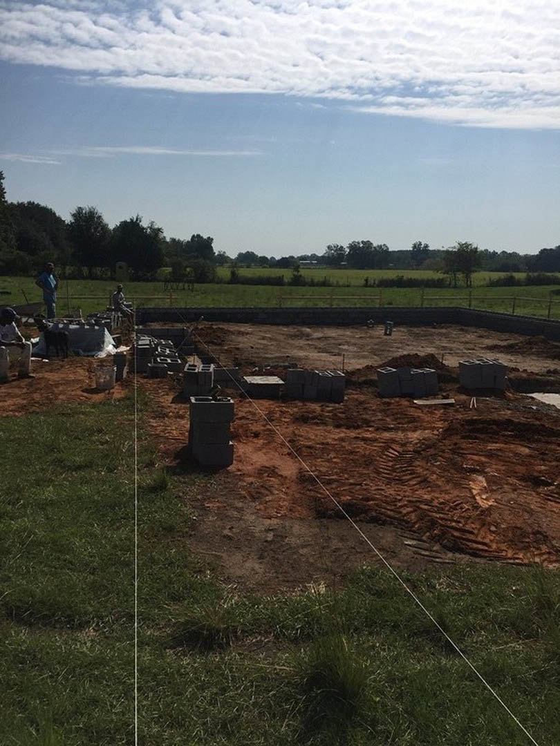 Partially built custom home on dirt lot with stacked black bricks, construction workers present, blue sky and scattered clouds overhead, grassy field and trees in background