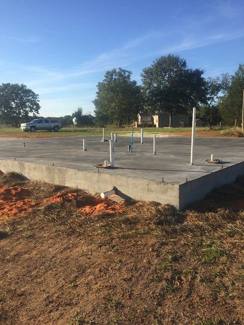 Concrete slab foundation with upright metal poles, nearby parked truck, scattered pipes, adjacent concrete wall, surrounding trees under blue sky