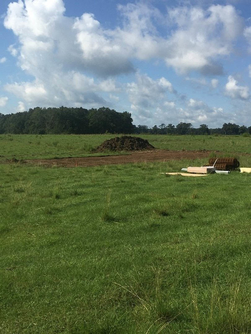 Grassy field bordered by a wooden fence, scattered trees in the distance, clouds overhead, pile of dirt near center, close-up roll of metal in foreground