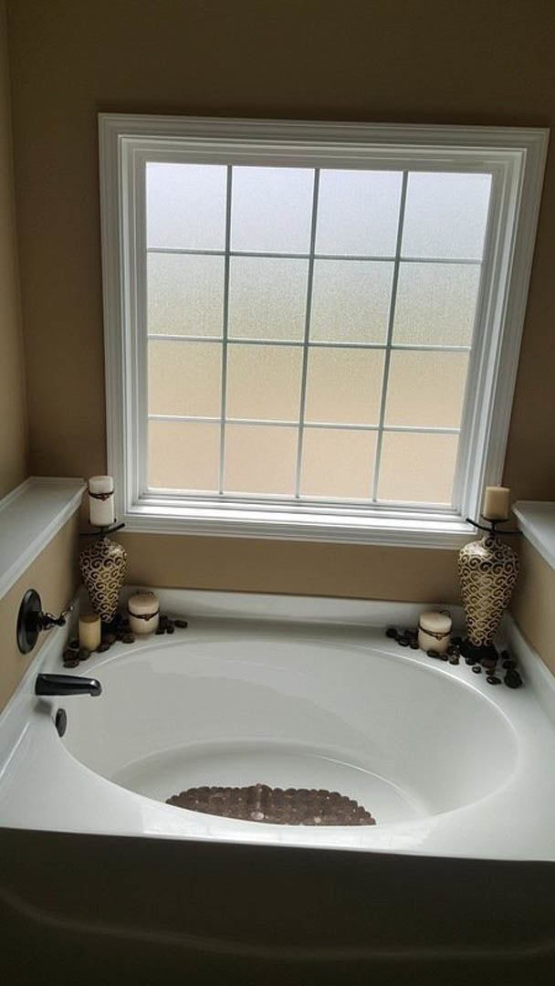 Freestanding bathtub with lit candles on the rim, large window with natural light, white tile walls, chrome faucet, and minimal decor