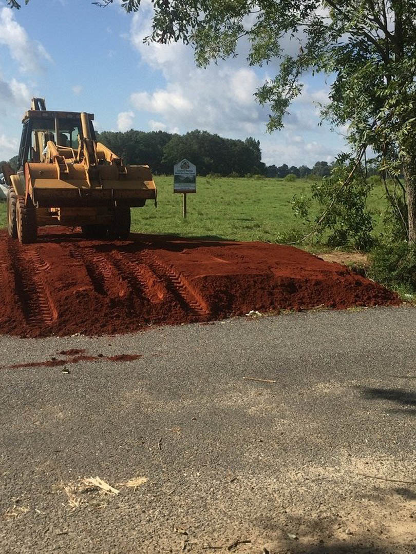 Yellow bulldozer parked atop a large dirt pile, tire tracks visible in soil, grassy area with construction sign, forested trees in background under partly cloudy sky