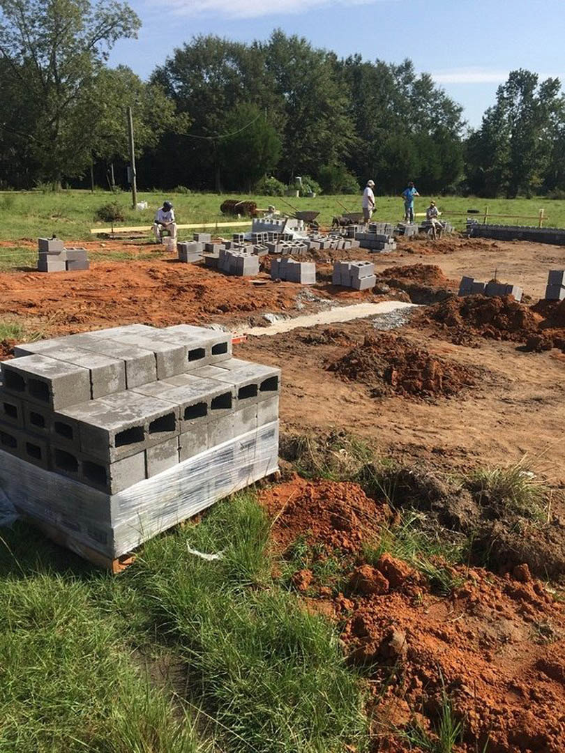 Workers assembling concrete block foundation on grassy lot, red dirt mound in foreground, trees lining background under open sky
