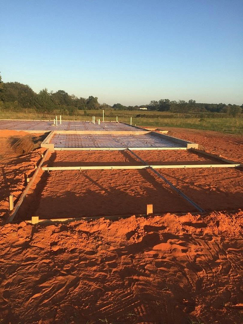 Concrete slab foundation with exposed rebar and metal frames on a dirt construction site under a partly cloudy blue sky