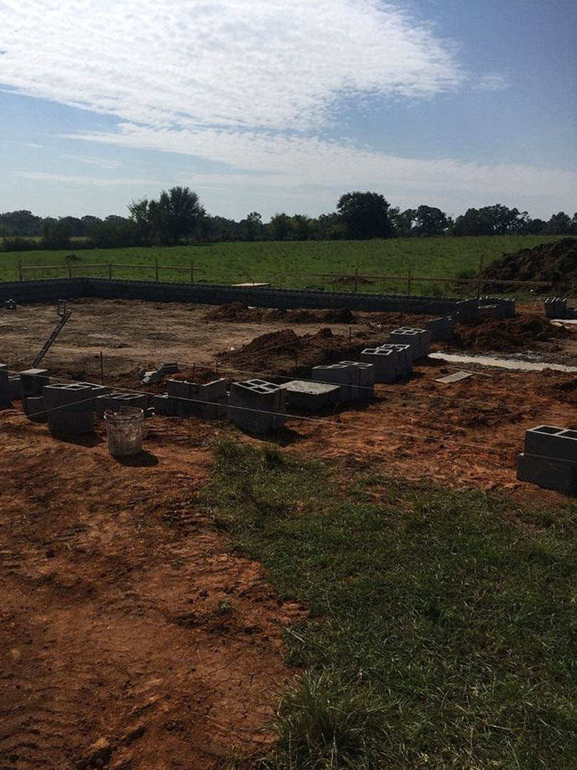 Framed construction site with exposed soil and grass, black foundation block, glass materials, open field, and distant trees under a partly cloudy blue sky