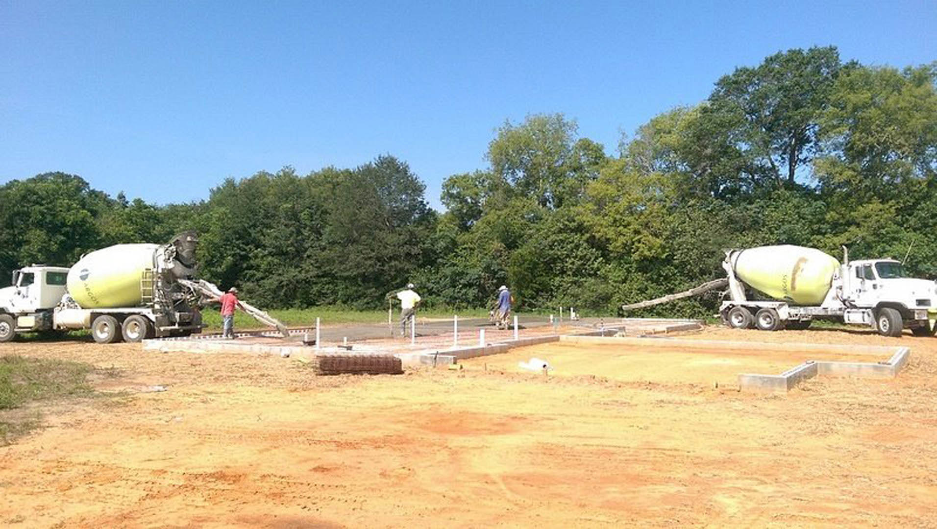 Construction site with workers, white cement mixer truck, dirt field, temporary fencing, and trees in the background