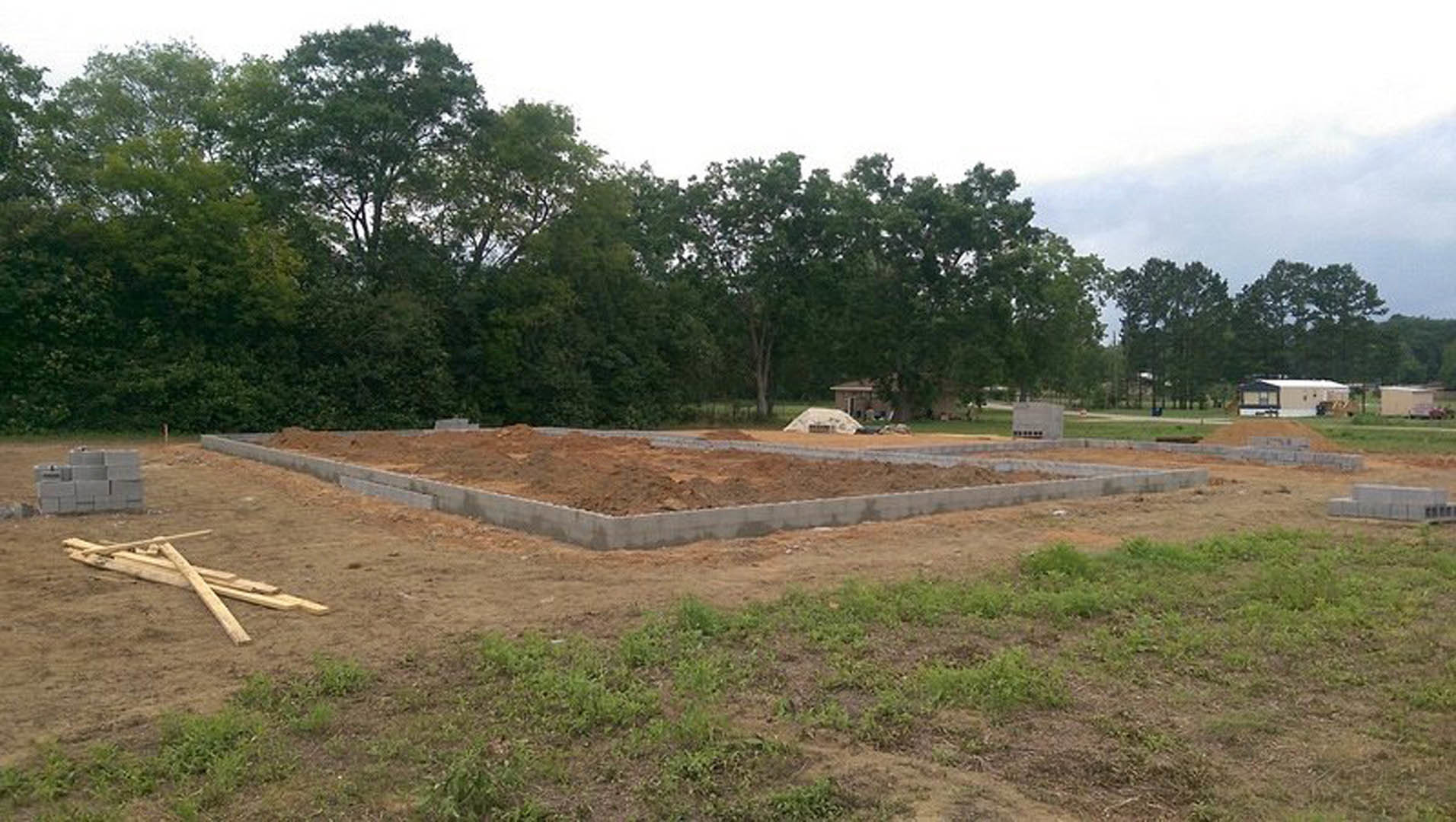 Concrete house foundation surrounded by dirt and grass, wooden planks stacked nearby, trees and cloudy sky in the background