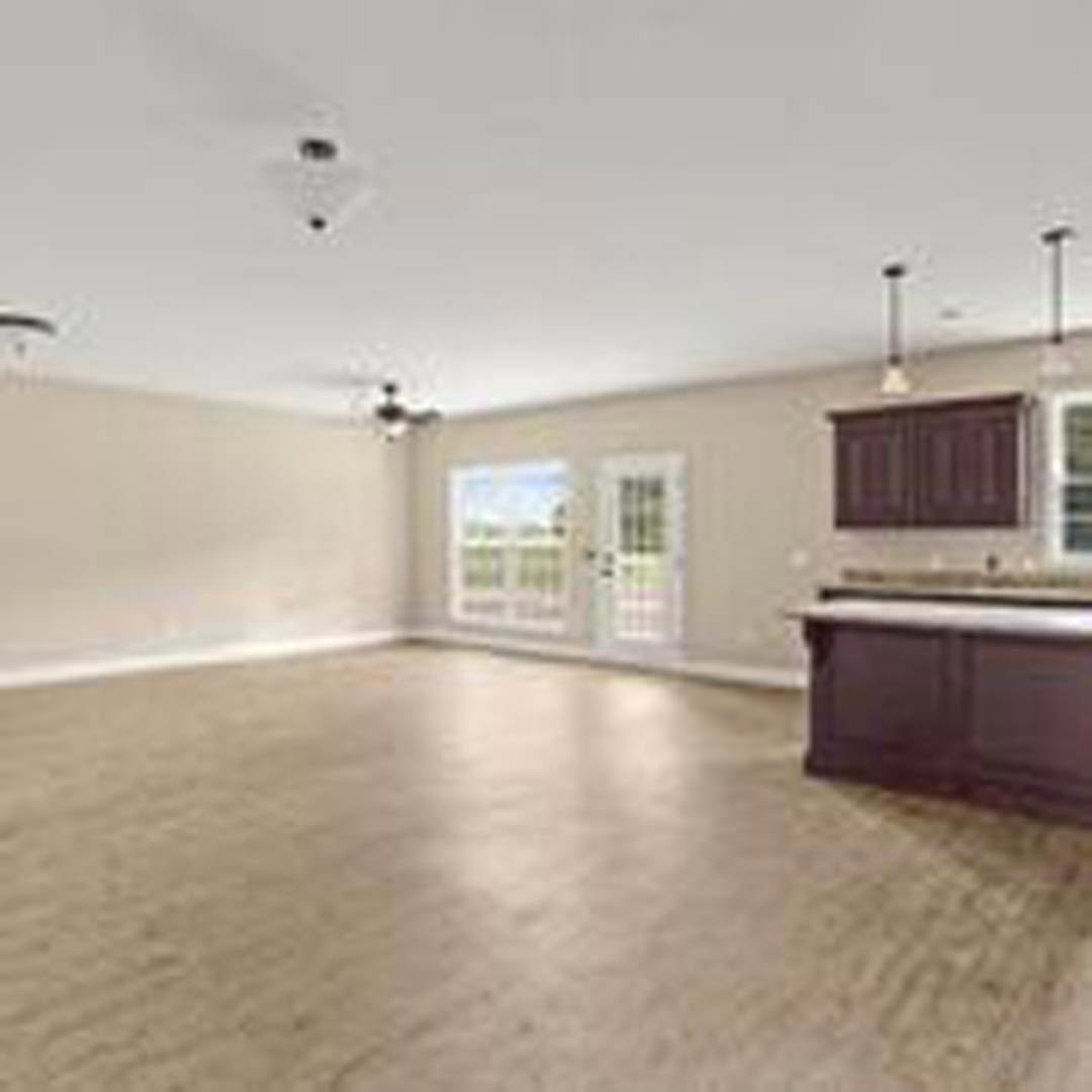 Spacious open room featuring a central kitchen island with light wood cabinetry, wide plank hardwood flooring, white walls, recessed lighting, and large windows providing natural