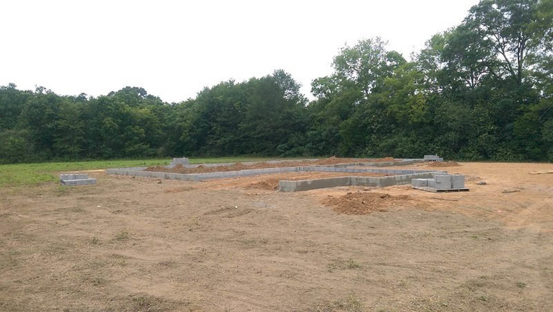 Concrete foundation set in a dirt lot, surrounded by grassy field and trees under open sky